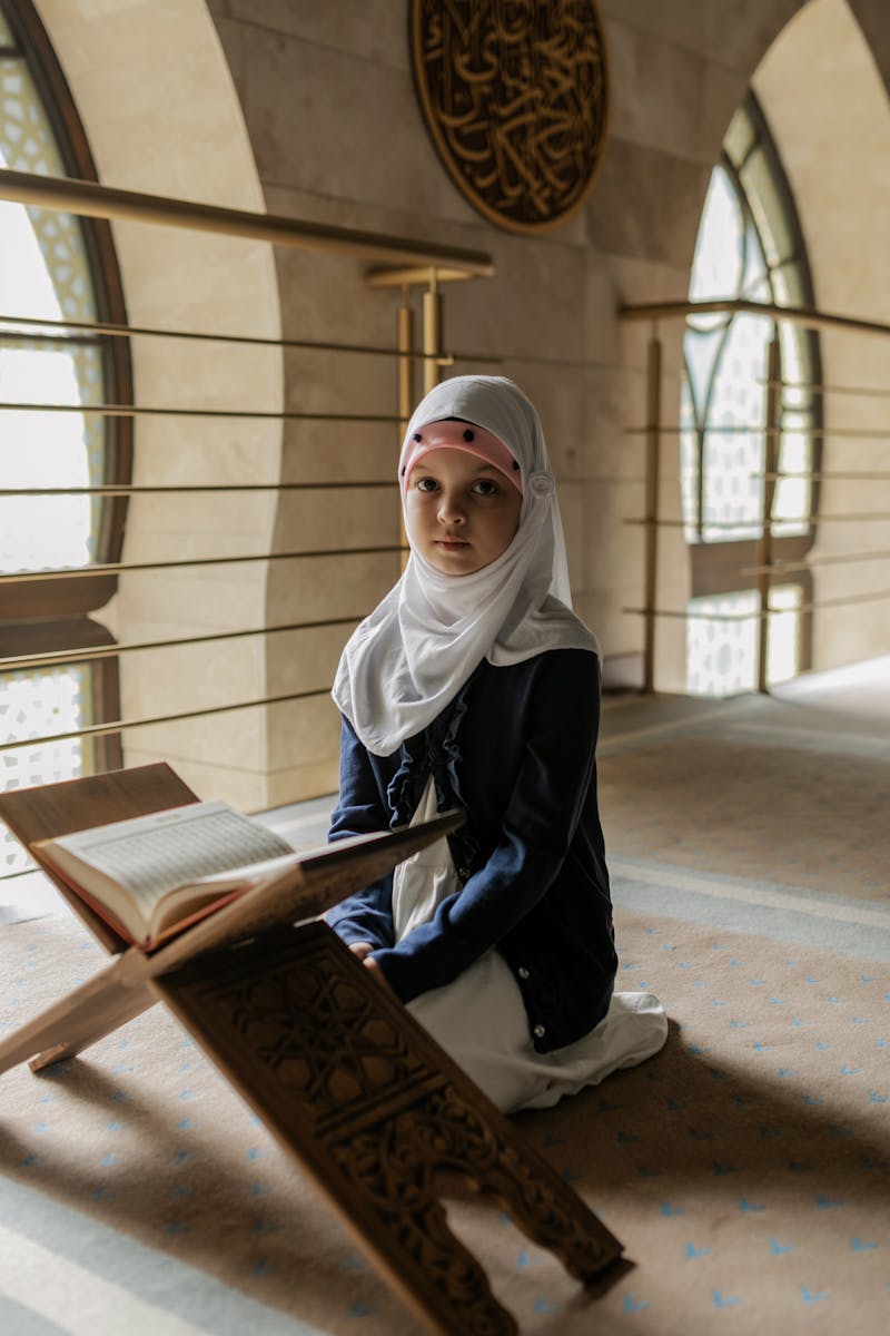 A young girl in traditional attire prays with the Quran inside a beautifully lit mosque, reflecting Muslim culture.